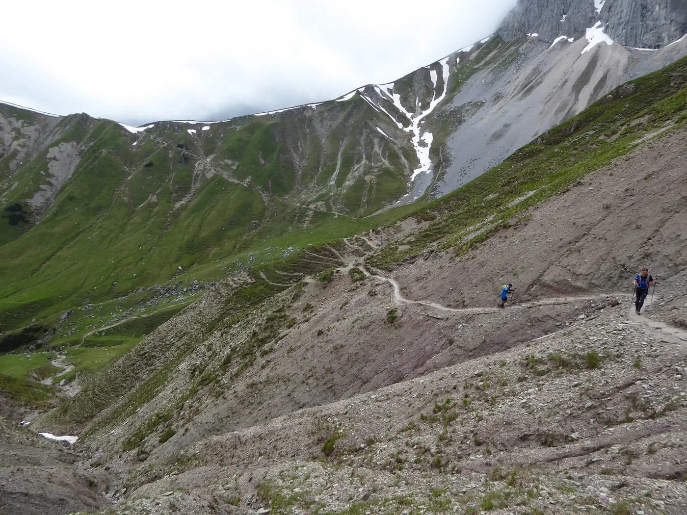 Runners on a steep rocky trail toward a snow-patched ridge